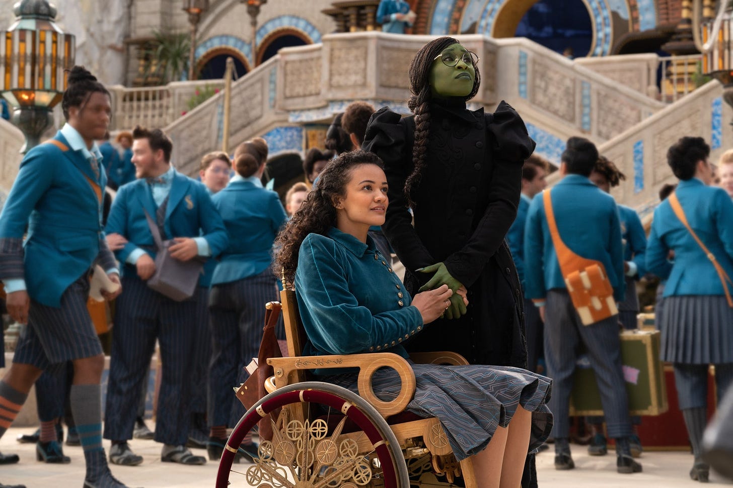 In a still from a movie, two characters are holding each other’s hands and looking in concerned amazement up at something out of frame. One has green skin and is wearing an elaborate black dress and large round glasses (played by Cynthia Erivo). The other is using an ornate manual wheelchair made of wood, gold, and maroon velvet. Behind them, students in blue private school uniforms are moving about.