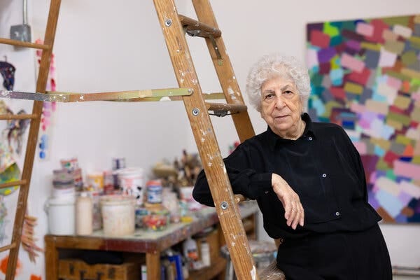 An older woman with white hair and a black outfit leans against a paint-splattered ladder. A table of paint and artistic materials sits in a corner behind her.