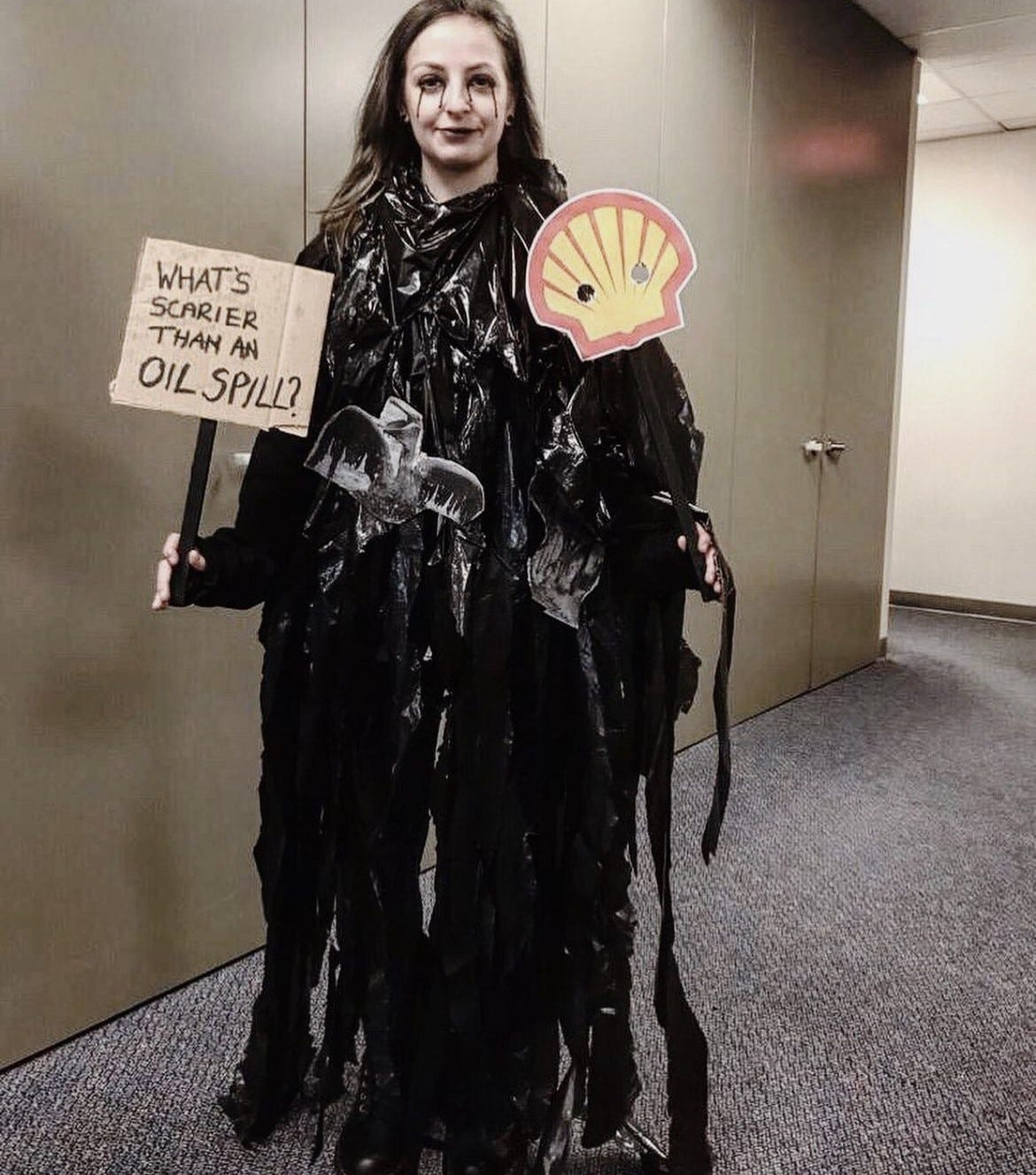Chelsea, a white woman, wears shredded bin bags to emulate an oil spill. Her blonde hair is sprayed black and she has black eye liner running from her eyes down her face. In her right hand she holds a sign reading 'what's scarier than an oil spill'. In the other hand she holds a face mask on a stick which is a shell logo with eye holes. On the black bags are black and white print outs of animals covered in oil.