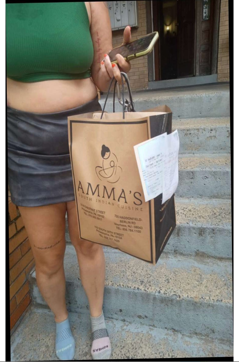 image of the author from the neck down, standing on a stoop holding a brown paper back from amma's south indian cuisine, wearing a forest green crop and faded black leatherette skirt, mismatched bombas sockettes.