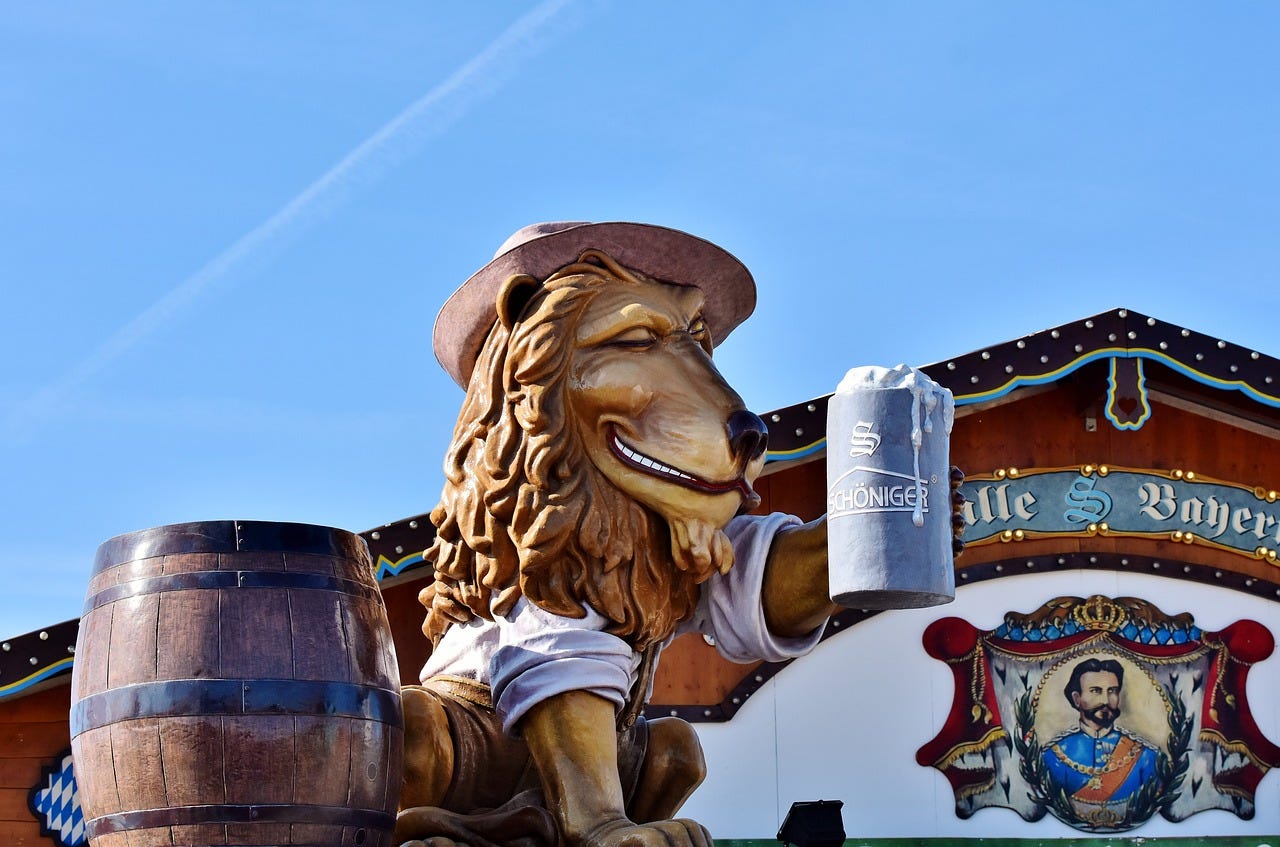 A large statue of a lion wearing a traditional Bavarian hat, holding a beer stein in one paw and sitting beside a wooden barrel, part of a festive Oktoberfest setup under a bright blue sky. A large statue of a lion wearing a traditional Bavarian hat, holding a beer stein in one paw and sitting beside a wooden barrel, part of a festive Oktoberfest setup under a bright blue sky.