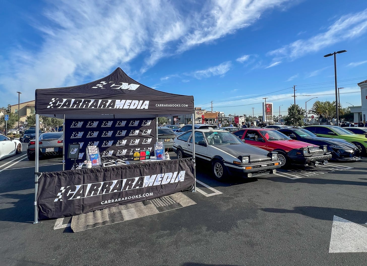 Photos of a Toyota Supra, Porsche Cayman GT4RS and Toyota AE86 parked next to the Carrara Media booth at a car show in Stanton, California.