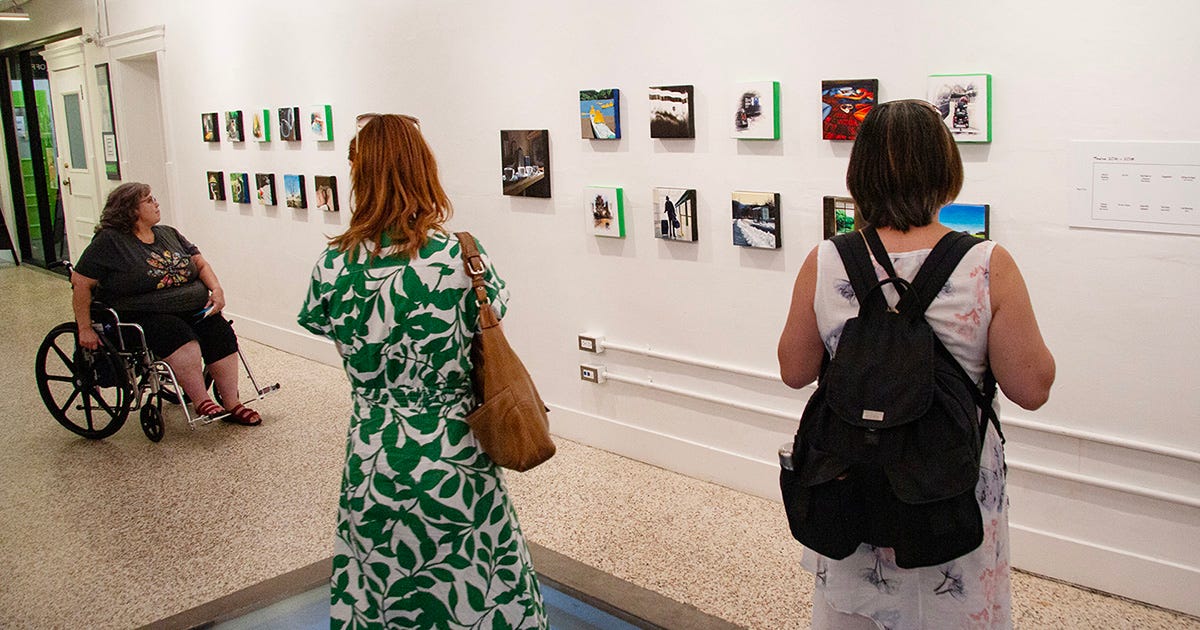 Three women look at a wall of small paintings in the gallery space. One is seated in a manual wheelchair and the other two stand with their backs to the viewer.