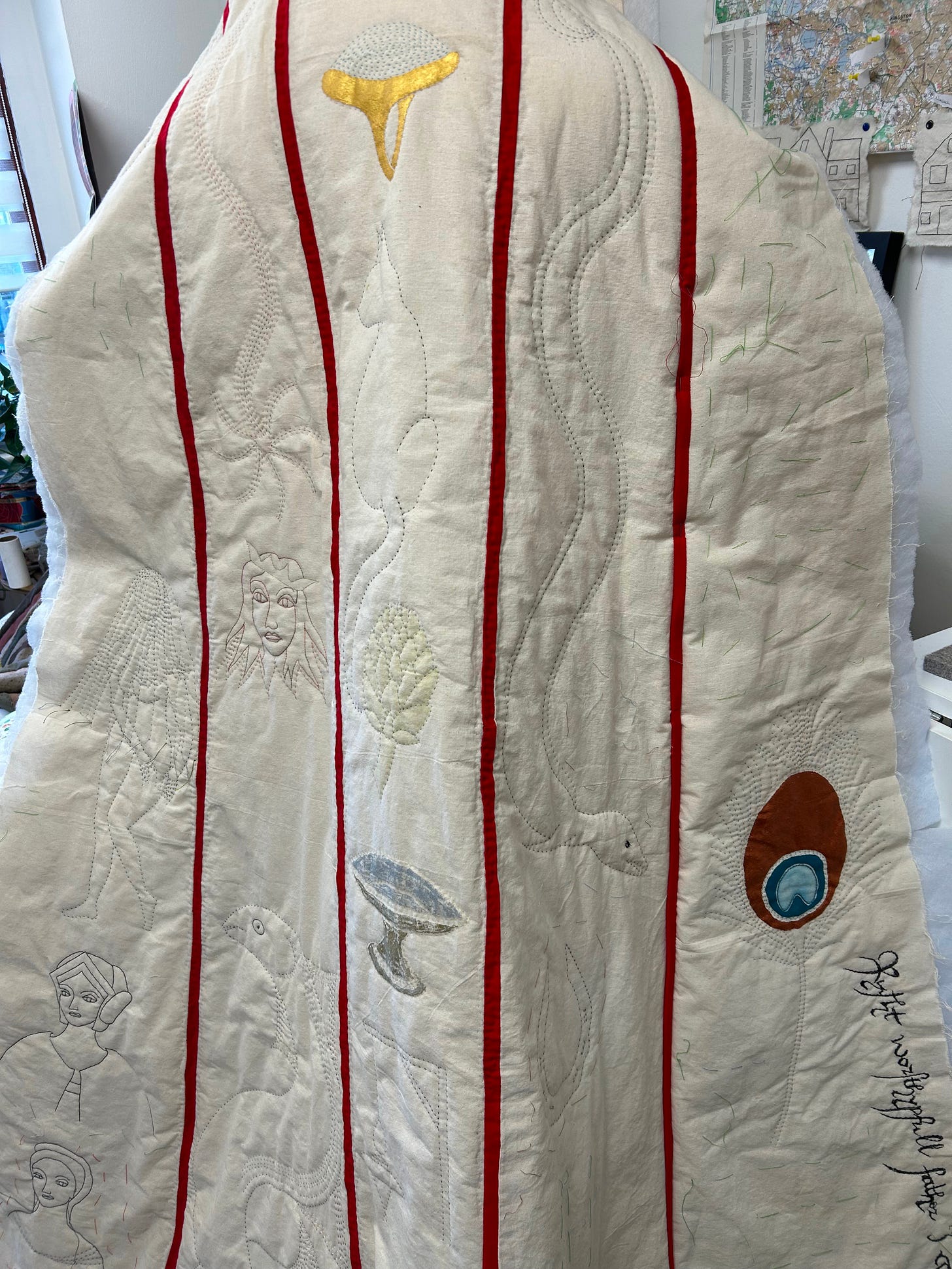 A partly made cloak, quilted and embroidered with various emblems: a gold and turquoise ring, a cat, an artichoke, women’s heads, a comet, an eel, some handwriting, a peacock feather. The cloaks i made of off white fabric, with red stripes joining each section.