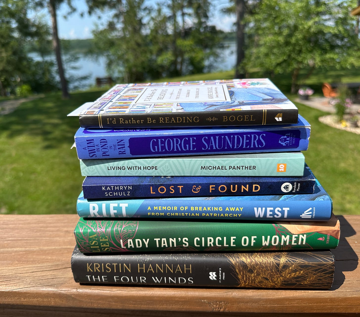 Stack of books with lake in background. Books: The Four Winds (Hannah), Lady Tan's Circle of Women (See), Rift (West), Lost & Found (Schulz), Living with Hope (Panther), A Swim in a Pond in the Rain (Saunders), I'd Rather Be Reading (Bogel)