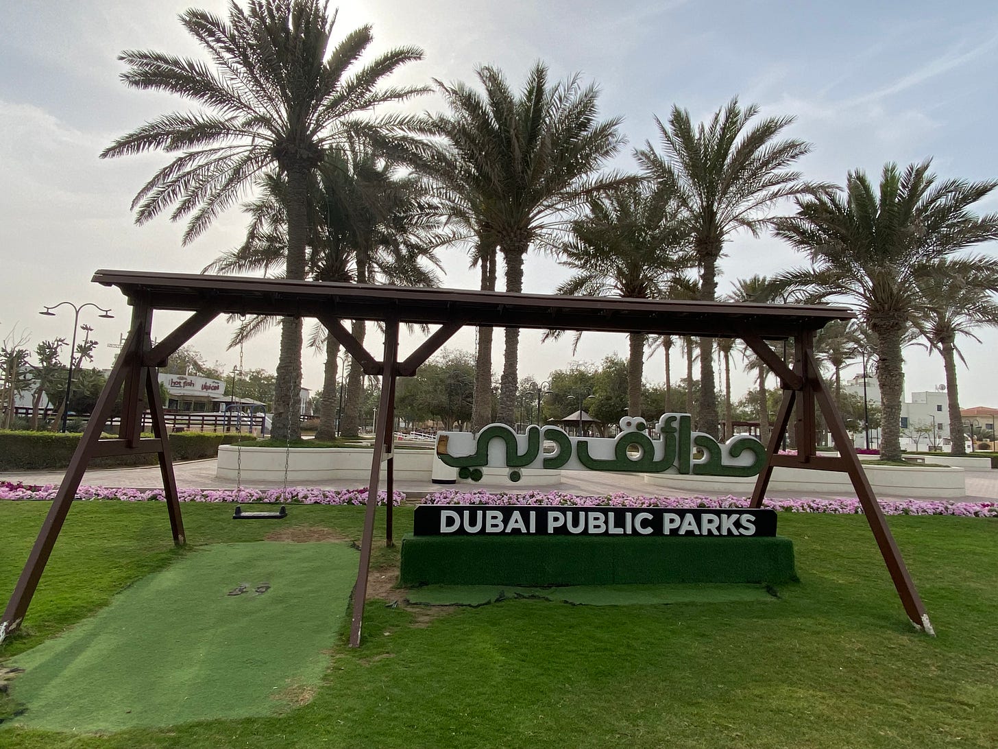 Photo of a park with a swing and palm trees in the background during mid-morning hours. There is a sign to the right of the swing that says "Dubai Public Parks" in both English and Arabic