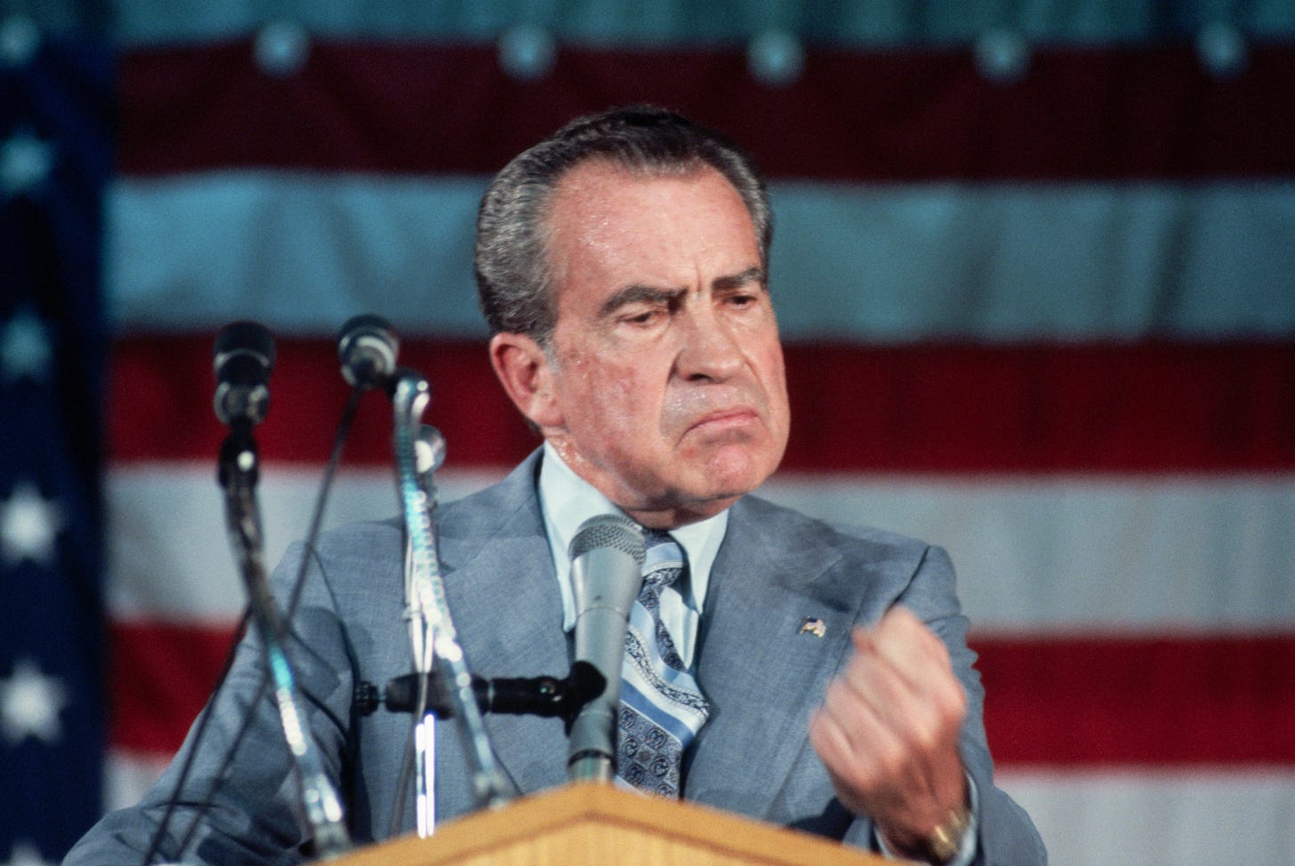 Richard Nixon, an older white man with swept back hair in a grey suit, stands behind a podium with microphones in front of an American flag.