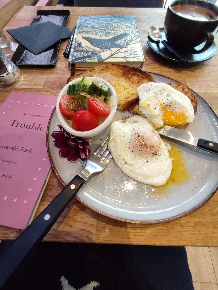 Table with breakfast of fried runny eggs, tomatoes, cucumbers, toast and a flower with knife and fork on a plate. to the left a copy of Trouble by Amanda Earl. above a journal with  black bird and a hot beverage in a cup