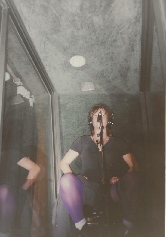 Woman in a recording booth singing into a mic. She has purple tights on and short hair