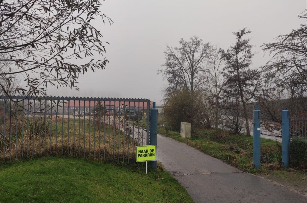 "Naar de parkrun" sign: "To the parkrun" on a metal fence, with a parking area beyond, just next to the start/finish.