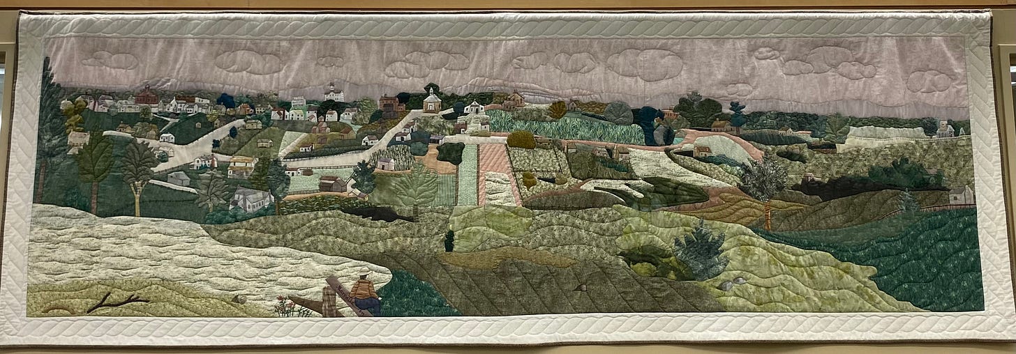 A quilt with buildings, grasslands, hills, houses, and shrubs all in varying shades of green and peach and mauve. 