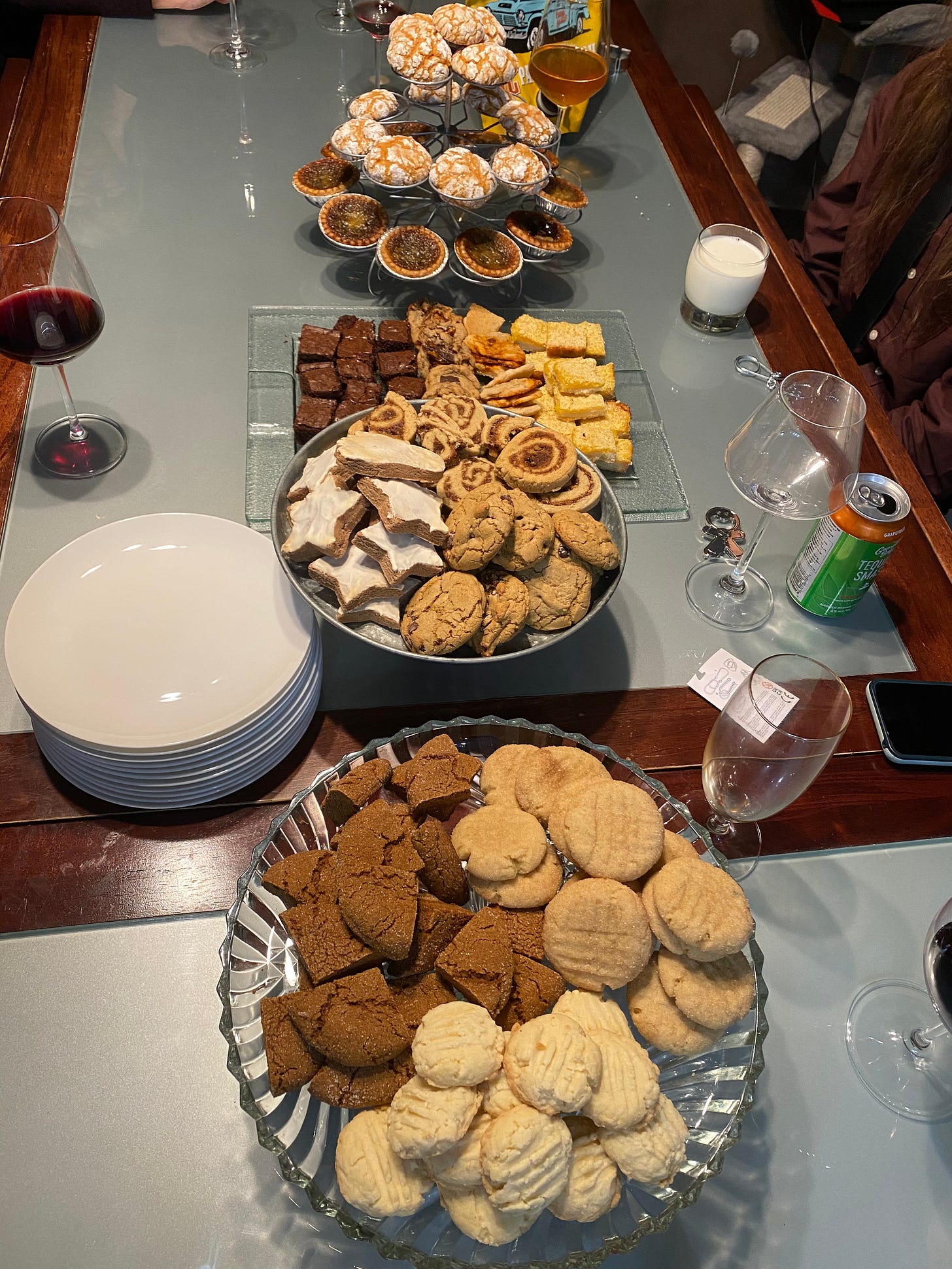 The cookies described below, set out on various plates and trays across a table with a scattering of empty and full wine glasses on it. A stack of plates is next to the central tray.