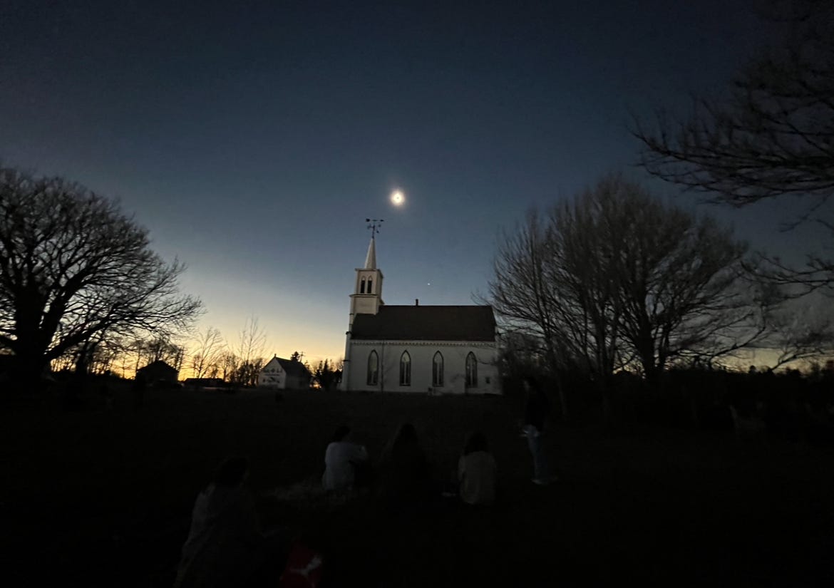 the solar eclipse in an eerily dark sky with an old white country church and skeletal trees in the foreground