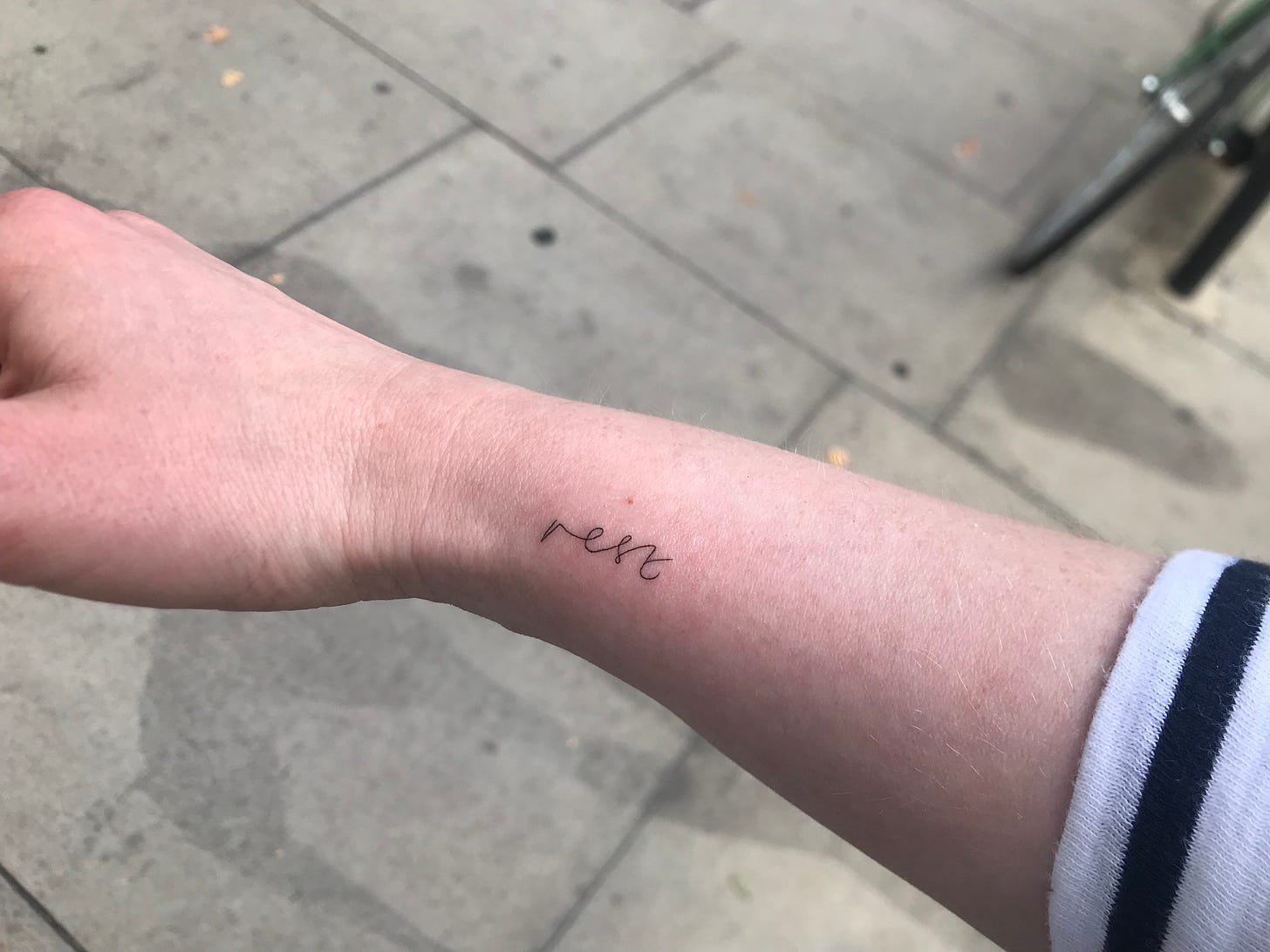 tattoo of "rest" in cursive script on a white woman's forearm, with pavement in background
