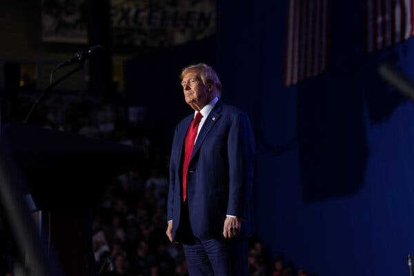 Donald Trump, wearing a blue suit and red tie, stands spotlighted onstage. An audience is visible behind him in the shadows.