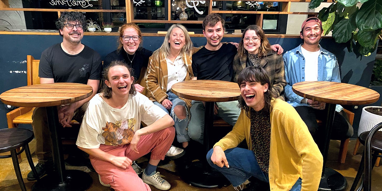 A group of friends smile for the camera sitting at three round coffee tables in a cafe. Two of friends crouch down in front with big smiles.