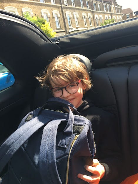 Joe, a cute boy aged about 10, in the back of an open top car, clutching his backpack on his lap and smiling a little nervously. 