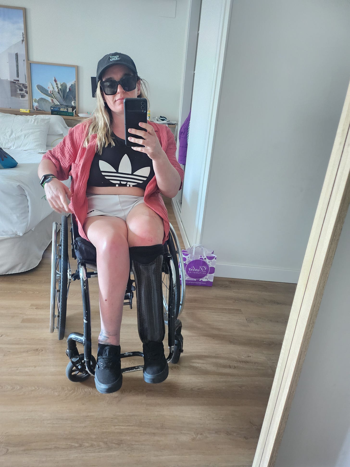 Taking a selfie in a mirror, I am in holiday mode. I am wearing a baseball cap, sunglasses, a pink shirt, a cropped Adidas Top, a pair of O'Neills shorts, and my fake leg is visible. 