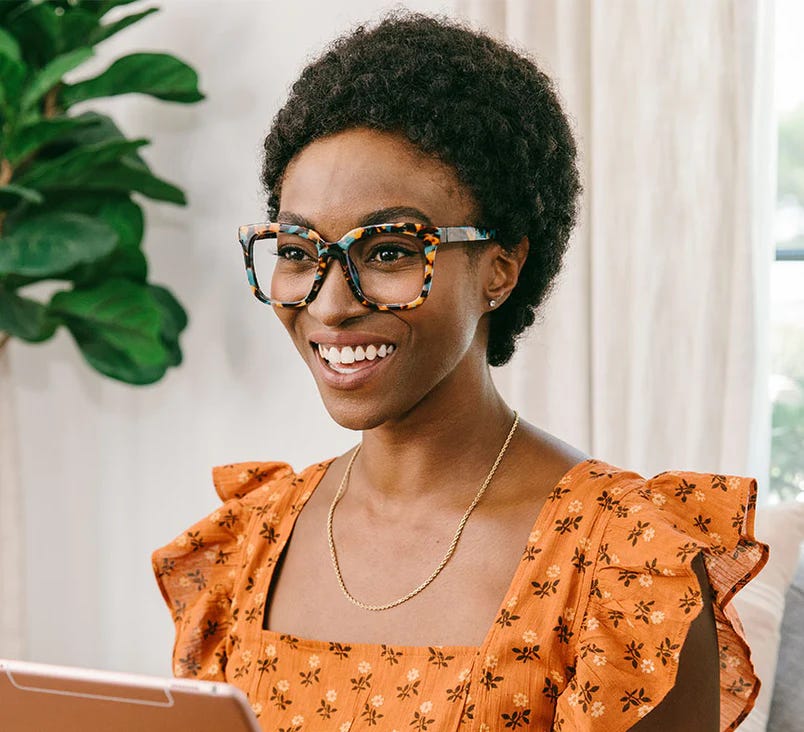 A Black woman smiling in a lovely orange dress wearing Peepers Next Level readers 