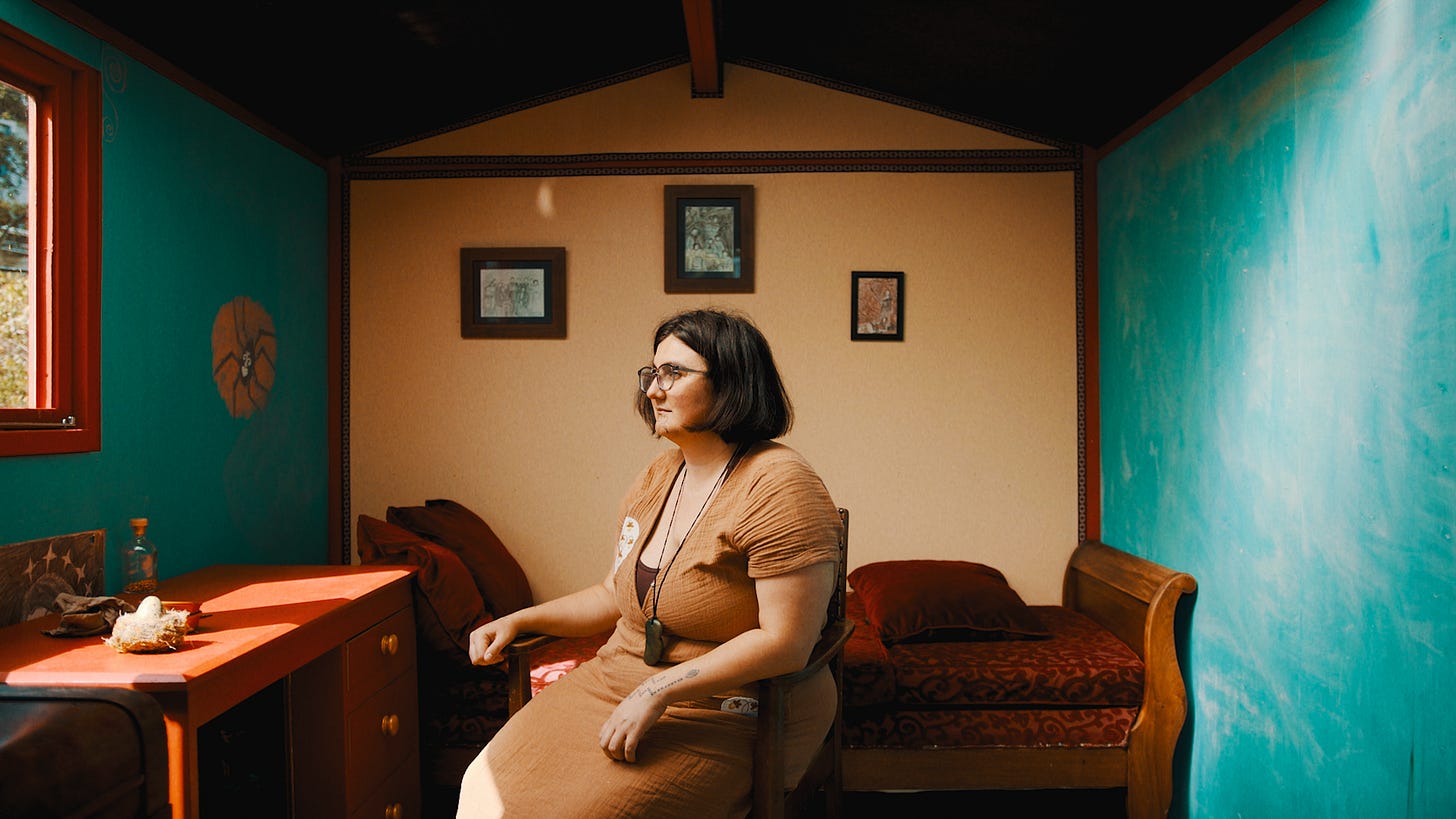 Ruby is a Māori woman with short brown hair wearing a tan-coloured dress and a pounamu around her neck. She sits in a chair in a blue and beige room. Photographer: Julie Zhu