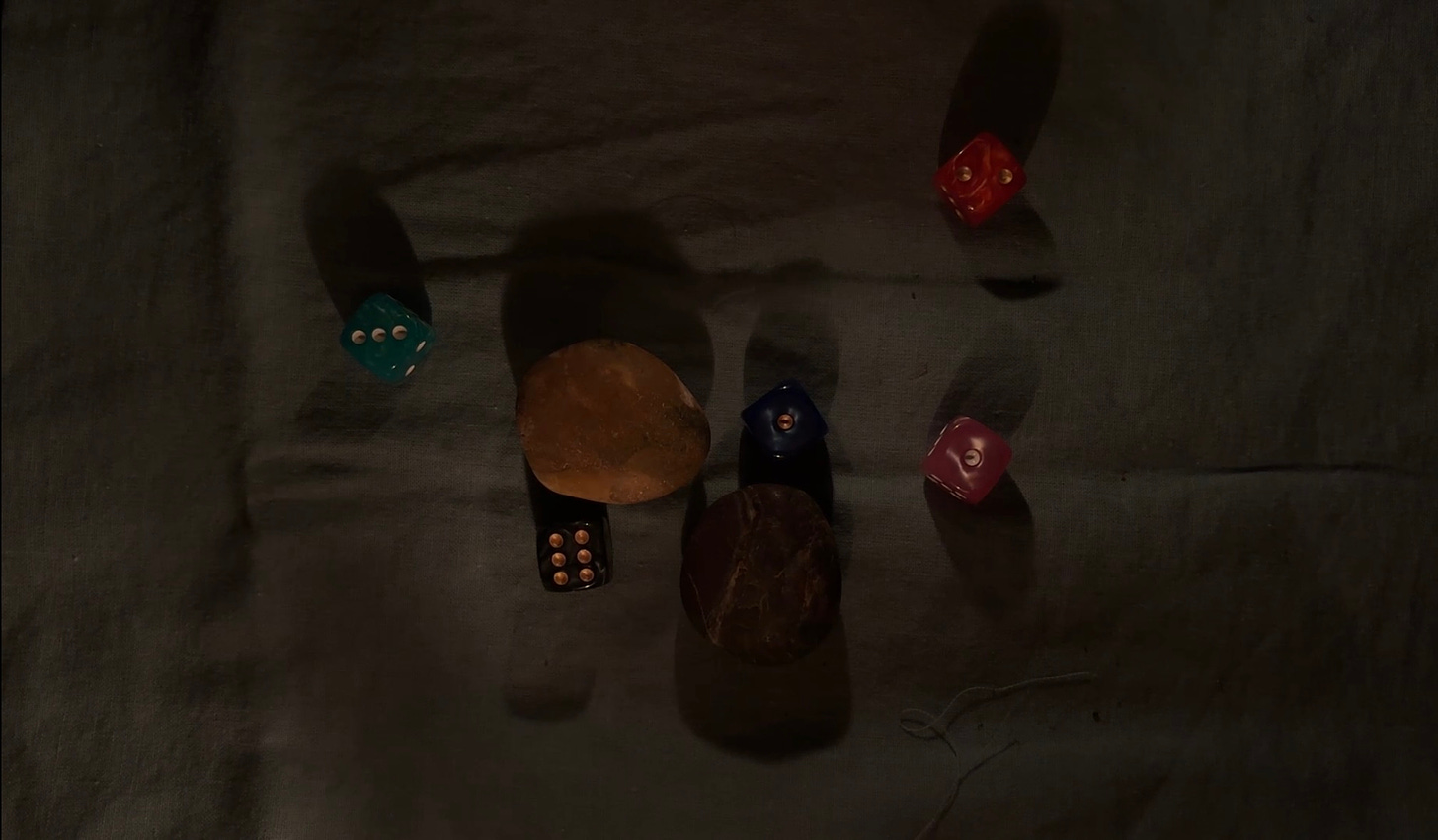 candlelit image of six colourful dice and two contrasting stones (light and dark) against a cloth background.