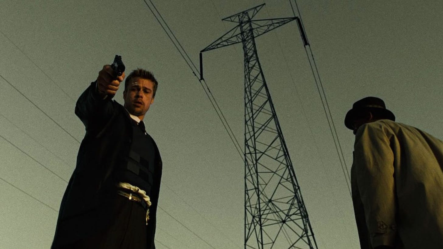 Fotograma de la película Seven. Brad Pitt con cara de confusión apunta a cámara con una pistola. Morgan Freeman está de espaldas.
