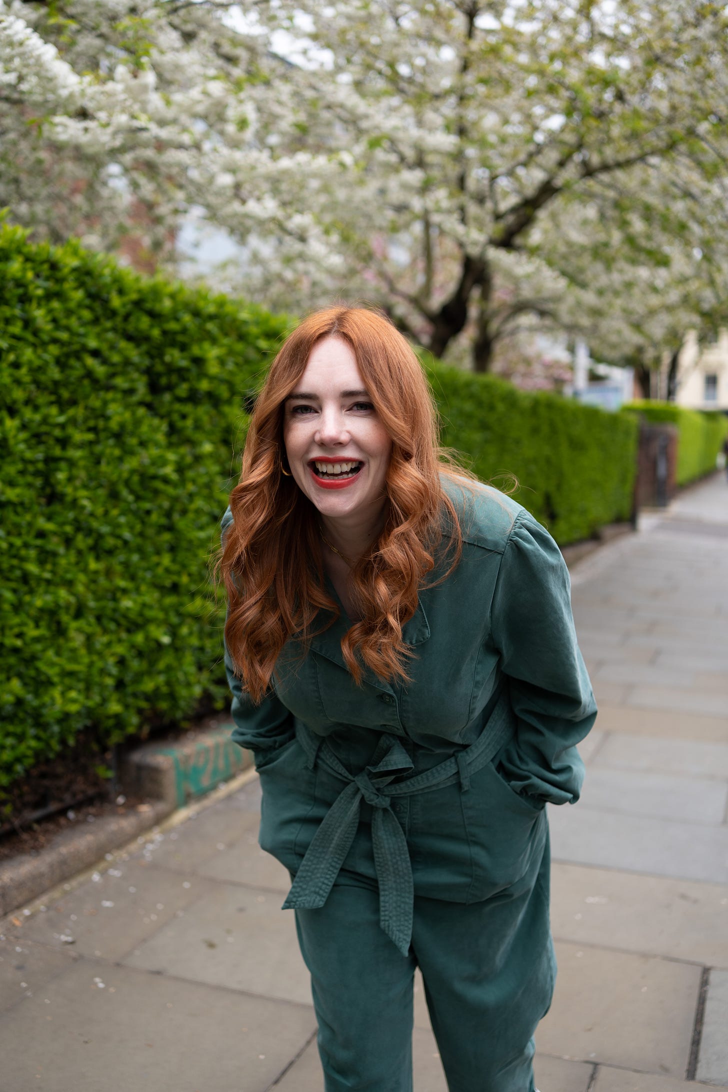Allie is leaning forward and laughing. She is standing on pavement next to a long green hedge with blossom trees in the background. She's wearing a green jumpsuit. 