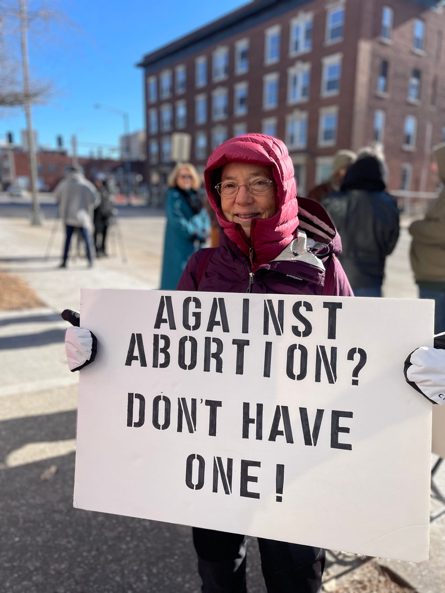 woman in hooded coat and mittens holds a large white homemade sign stating in bold lettering: "Against Abortion? Don't Have One!"