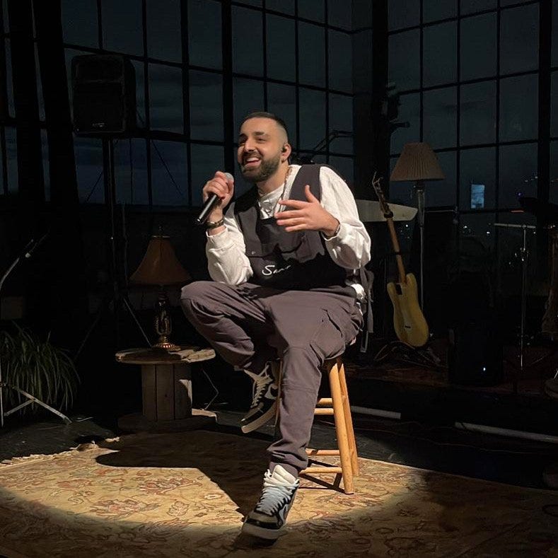 Photo of Samer Ajaj sitting on a stool smiling with a microphone in hand.