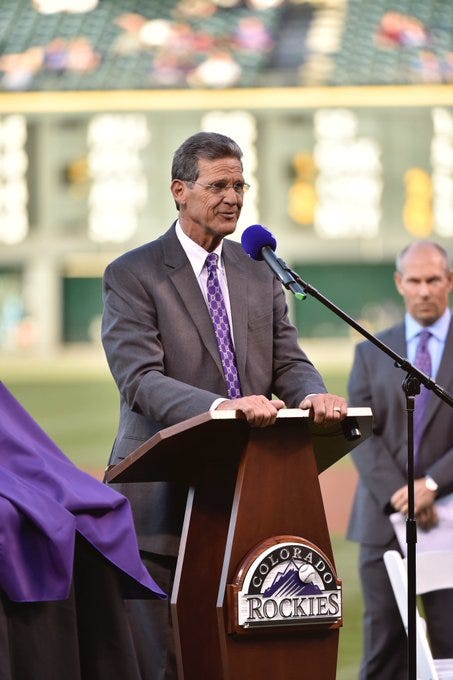 George Frazier, wearing a grey suit, purple tie, and white shirt, stands at a podium with the Colorado Rockies logo. 