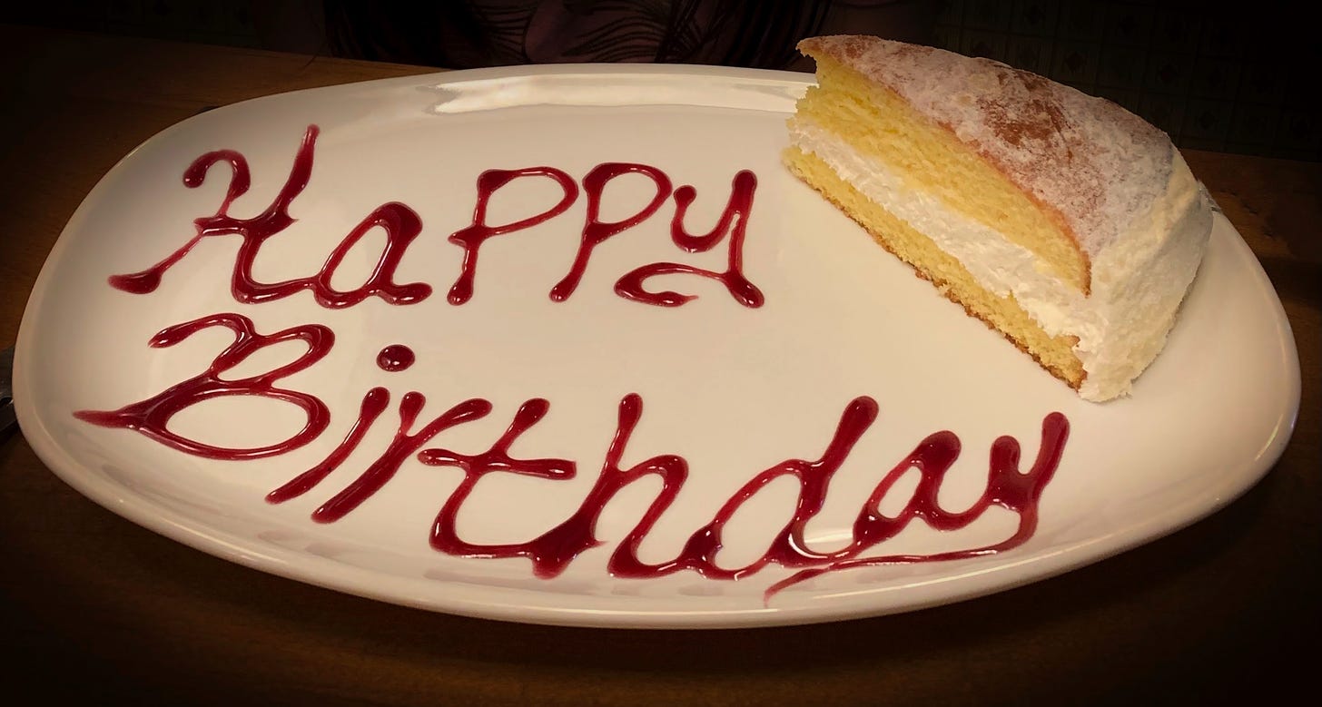 A white plate with Happy Birthday written on it with red icing and a slice of yellow cake with white icing in the upper right corner of the plate