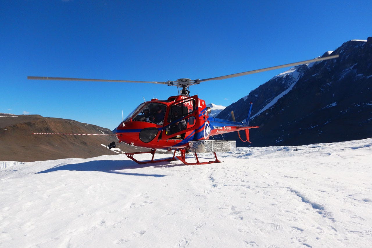 A red helicopter on the ground. The ground is icy and mountains are visible in the background. The tail of the helicopter has the words National Science Foundation.