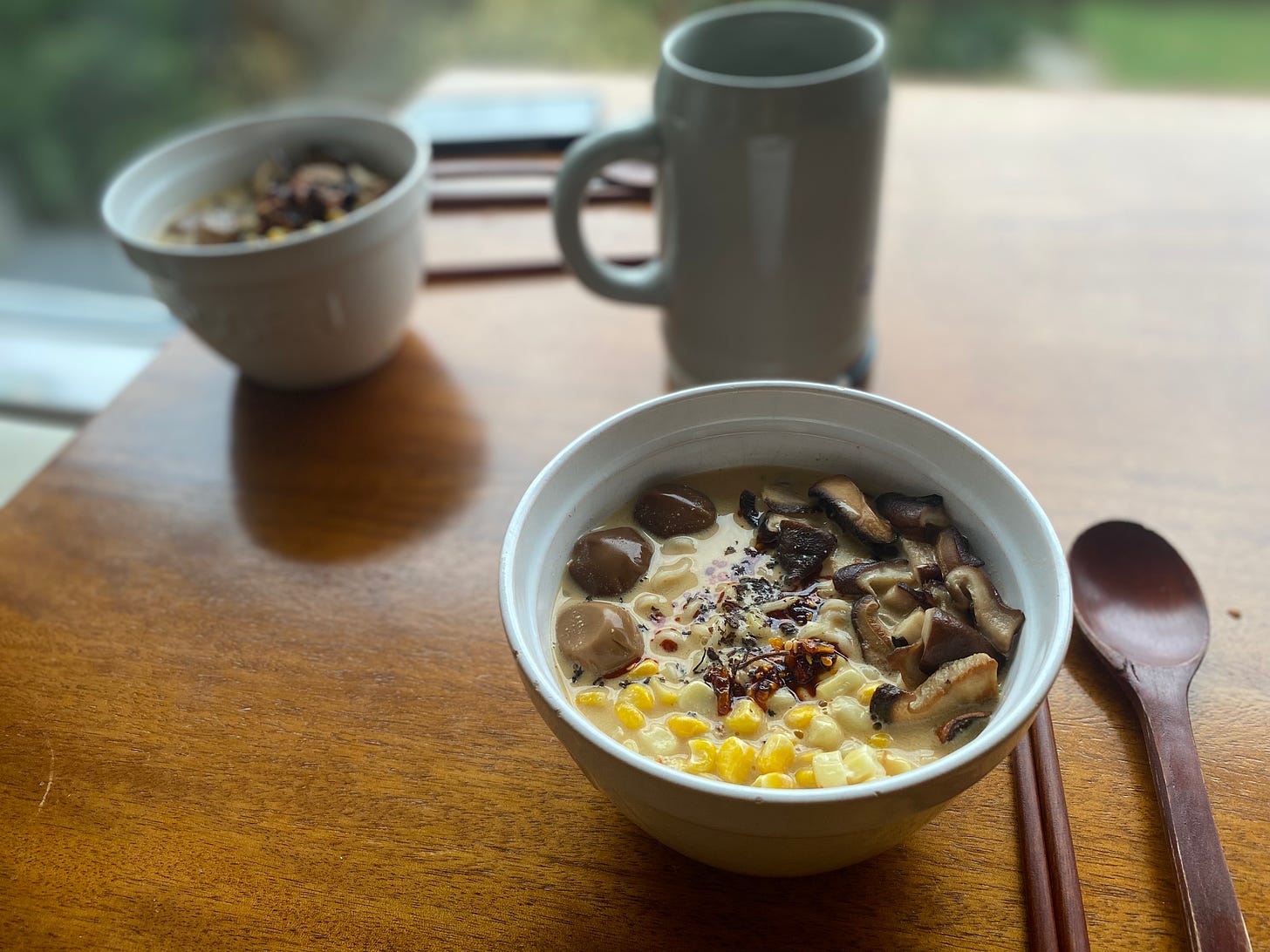 Two white bowls of ramen with the toppings described above: fried shiitakes, kernels of corn, and three marinated quail eggs, plus a drizzle of chili crisp. In the background is a large ceramic beer stein.
