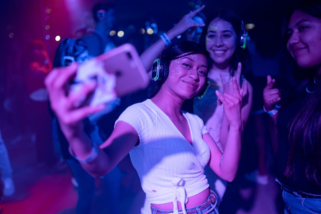 Free Young adults enjoying a silent disco party, capturing selfies amidst colorful lights. Stock Photo