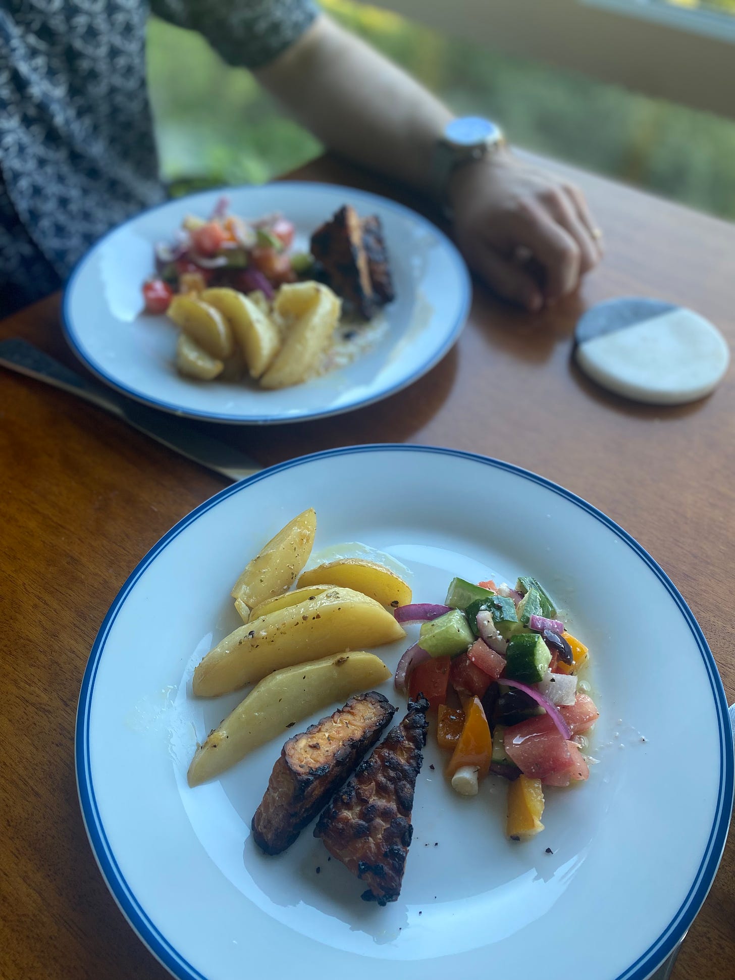 On a white plate with a blue rim, a few wedges of lemon potatoes and two triangles of blackened tempeh next to a small scoop of greek salad. Jeff's plate with the same is in the background, his hand resting next to the plate.