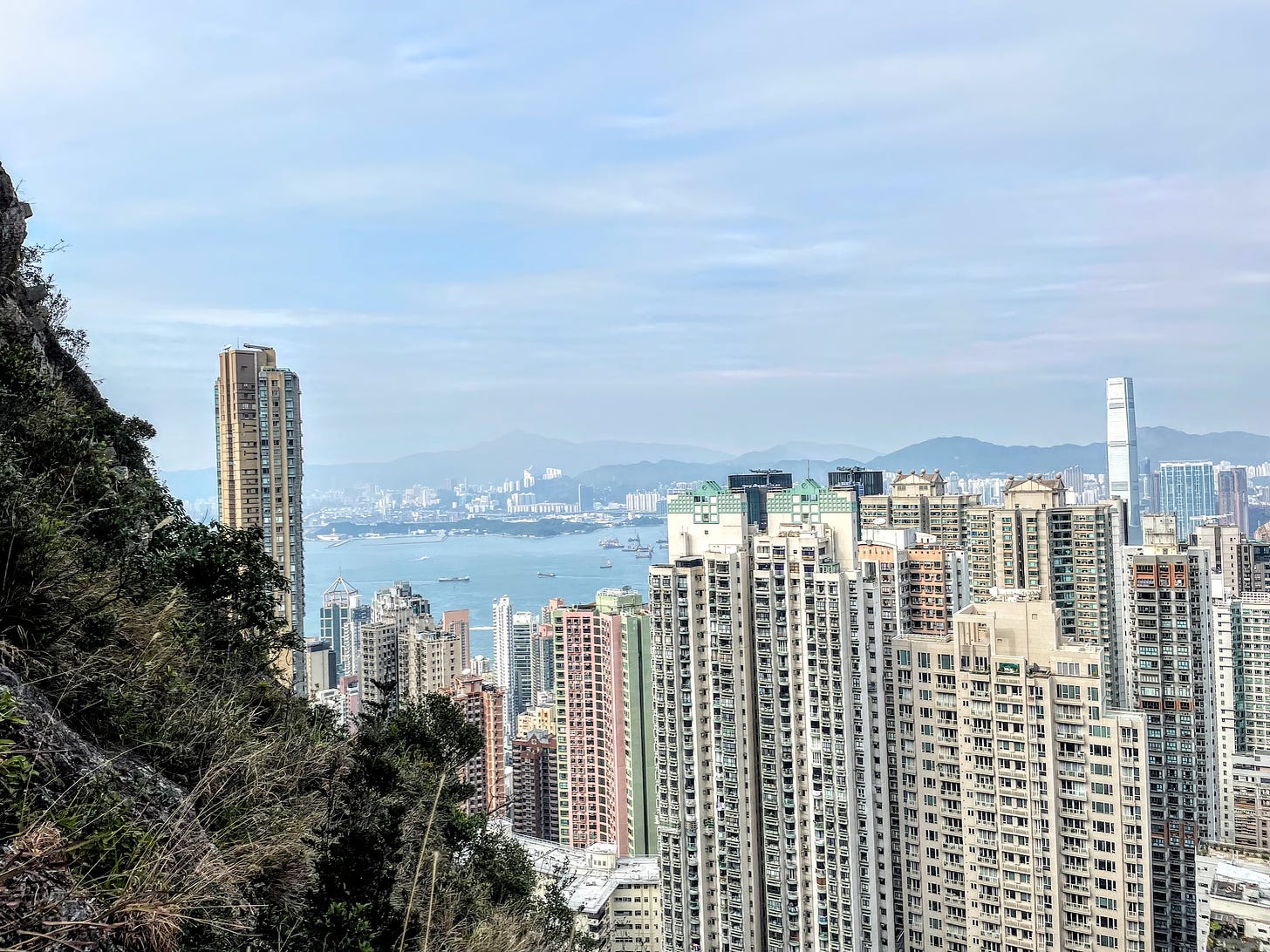 Hong Kong skyline from the side of a steep hill — many tall, colorful apartment buildings set before a large, blue bay, with rolling hills in the distance