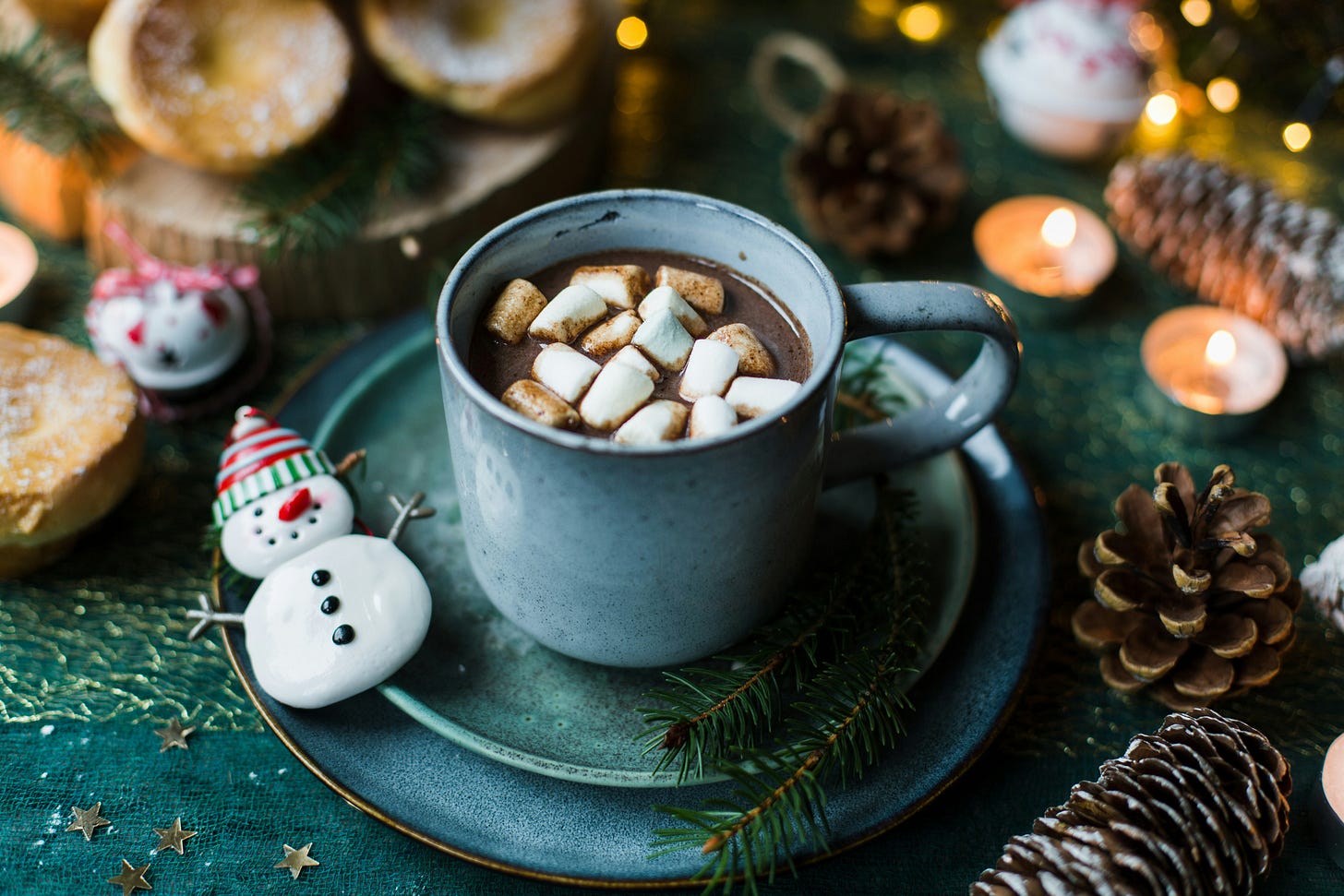 blue cup and saucer filled with hot chocolate and marshmallows with a snowman cookie on the rim of the saucer