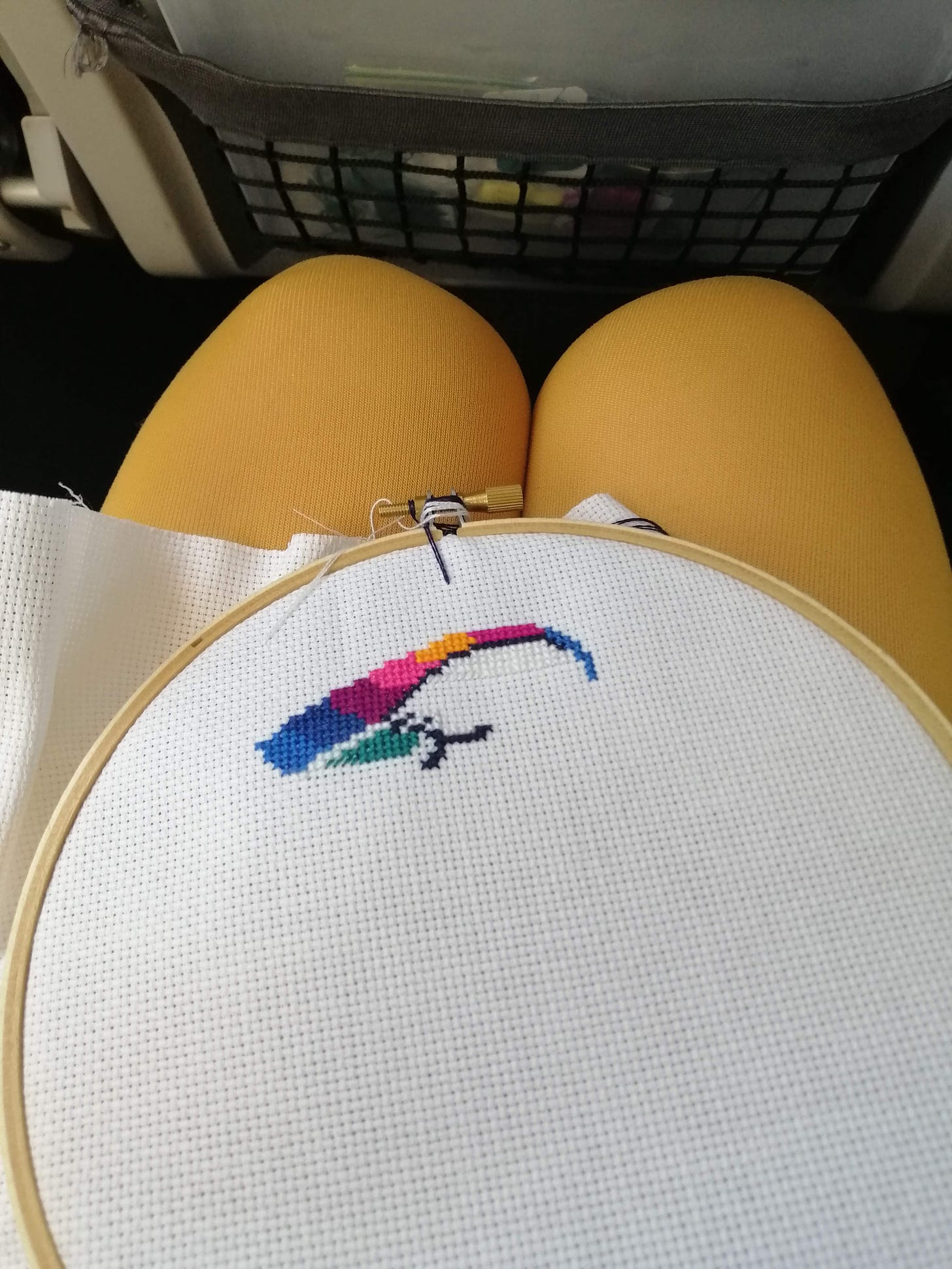 Photo taken inside an airplane showing my lap with a cross stitch project on it and stitching supplies in the seat pocket