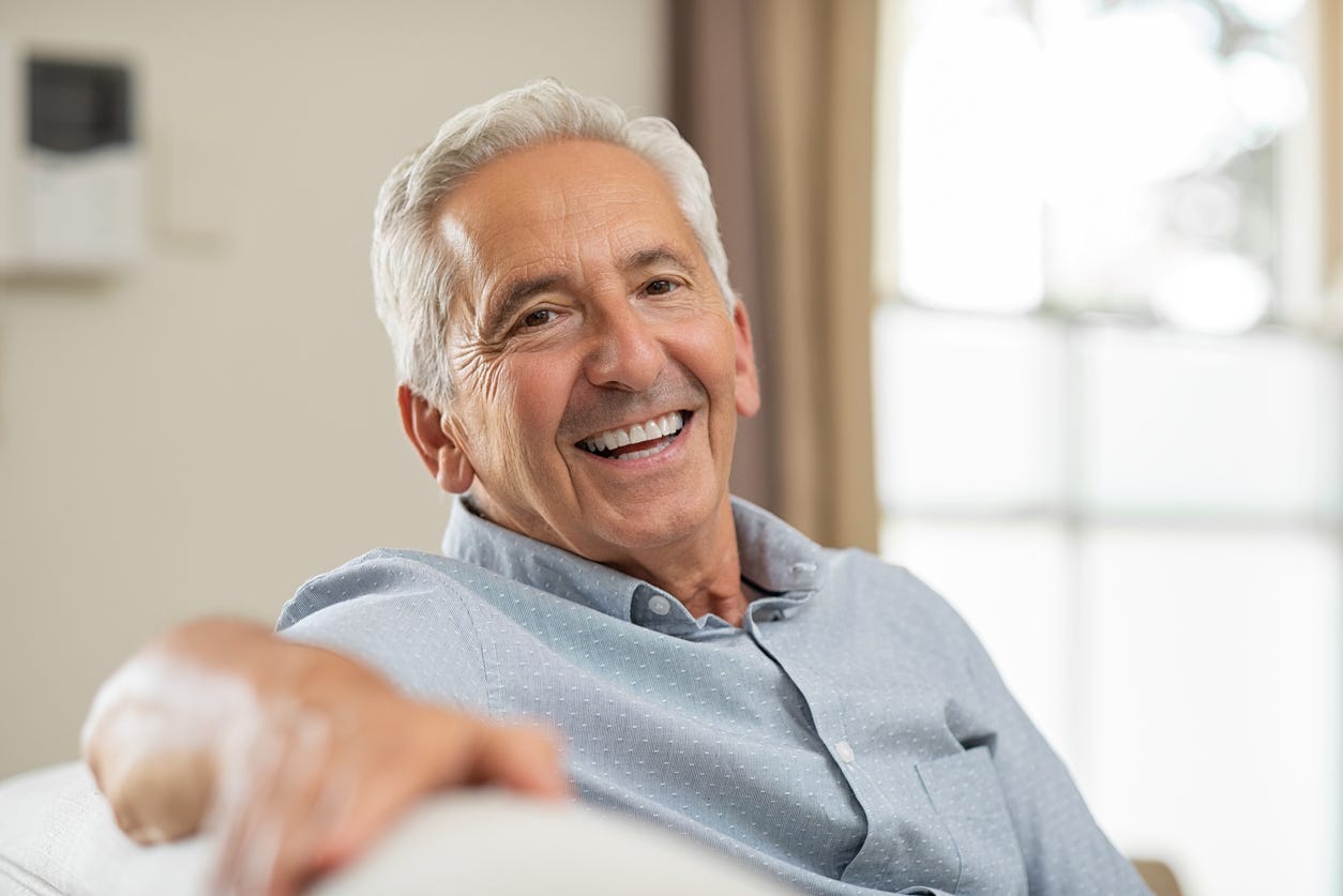 Smiling, older while man without glasses and wirh full head of gray hair, facing camera and extending right arm across top of couch.