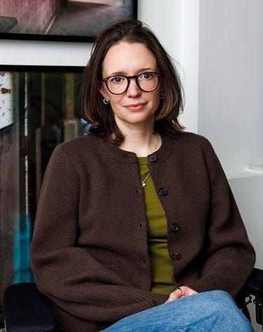 Jessica Slice with straight short brown hair, wearing brown rimmed glasses and a dark brown cardigan over a green shirt and jeans