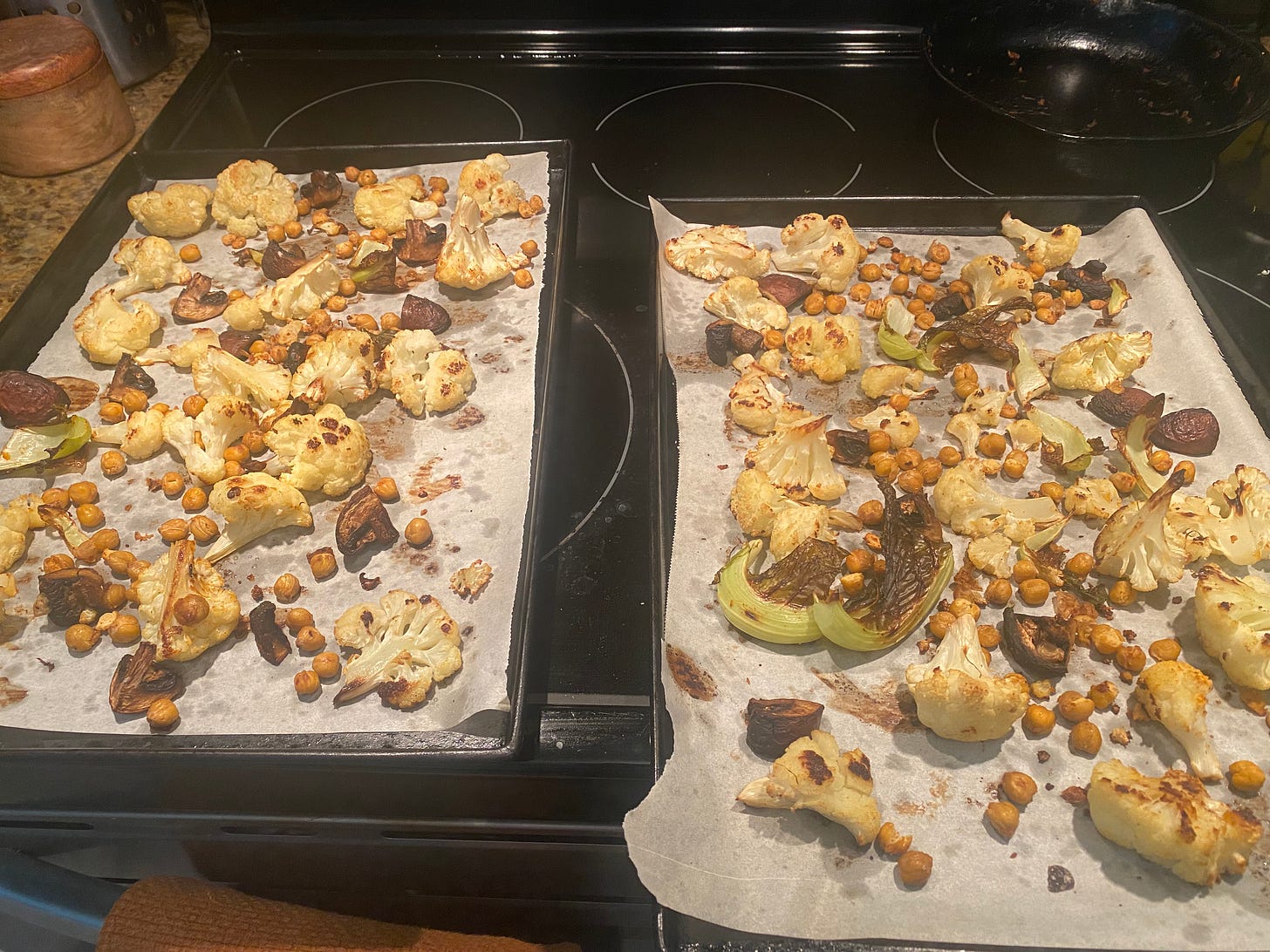 Two sheet pans lined with parchment, resting on top of the stove. Each has browned cauliflower and mushrooms, and crispy chickpeas.