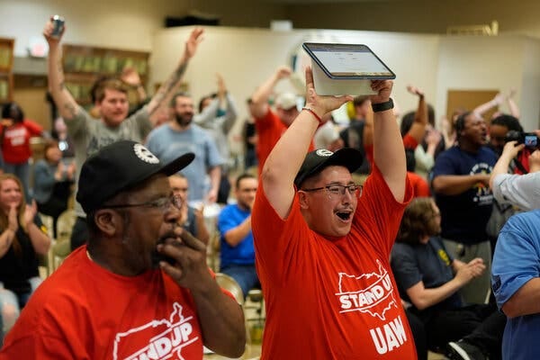 In a crowded room, men in T-shirts, some that read “Stand Up UAW,” cheer and hold up their hands.