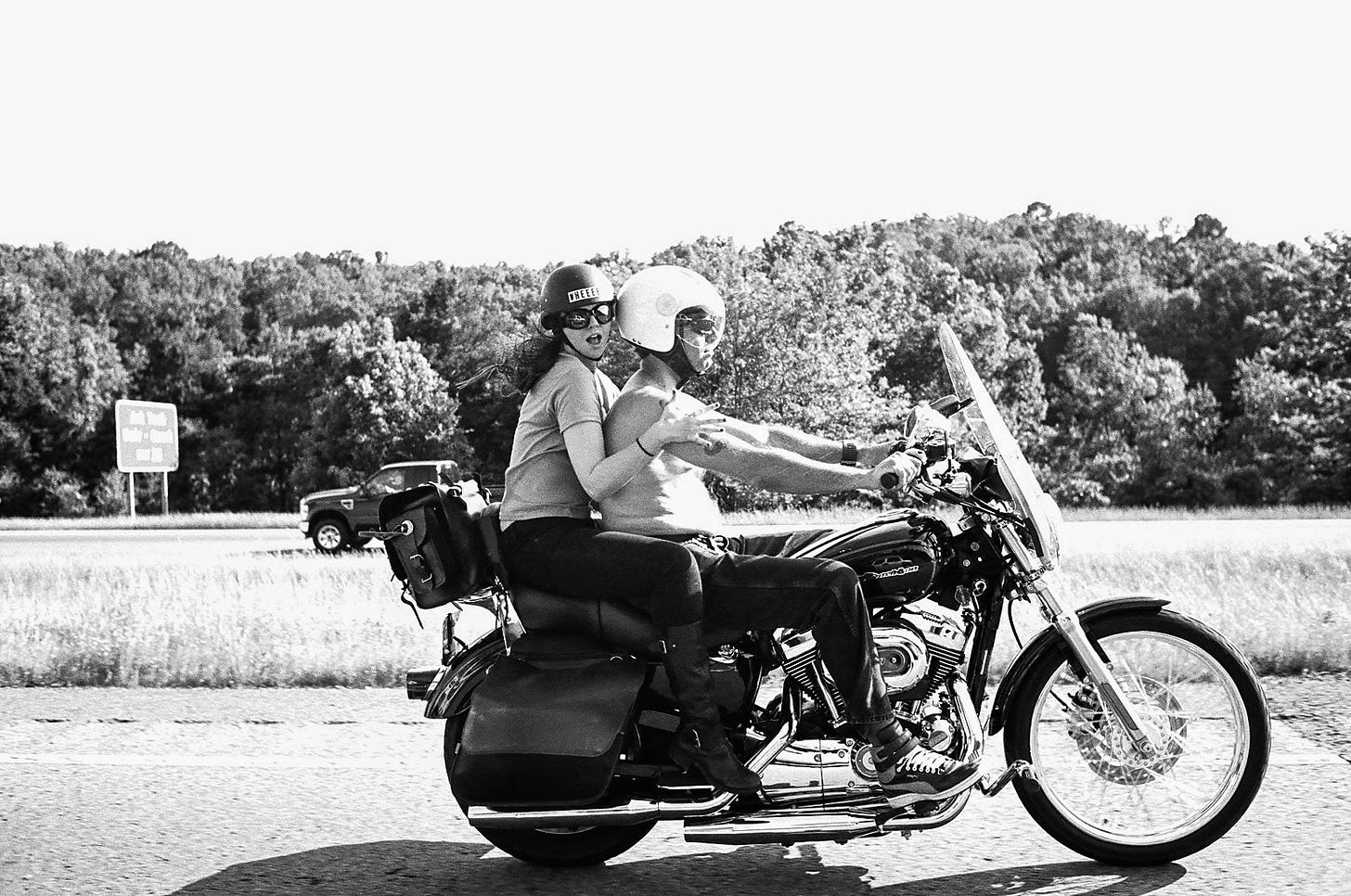 Derrick driving his motorcycle with Amber on the back. The photo is taken from the side of the bike. Derrick is shirtless, looking ahead at the road. Amber holds onto Derrick but is looking to the camera. They both wear helmets. Amber's helmet has a sticker on it that says "WHEEEE." 