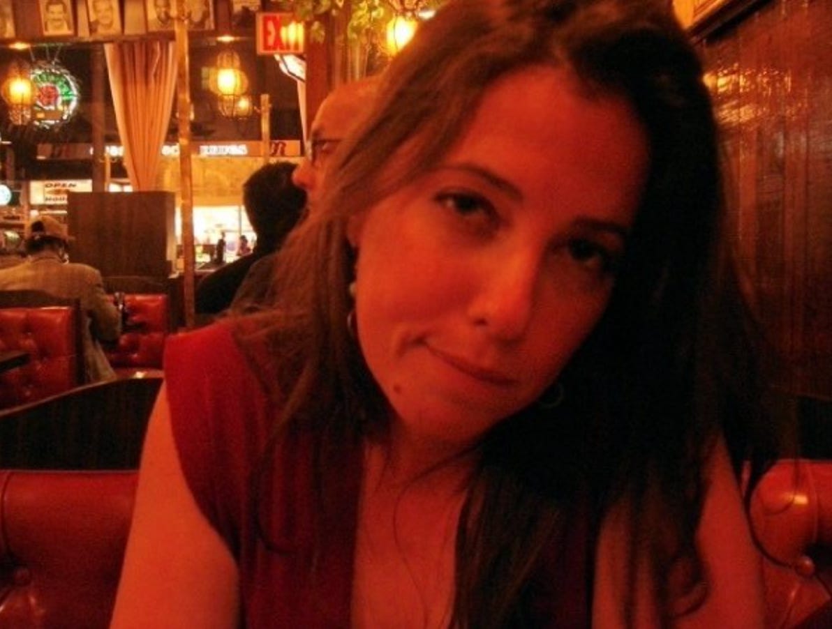 A woman in a red dress, the author, Holly Starley, leans on a table in a dimly lit booth, holding the camera's gaze, nearly biting her lower lip.