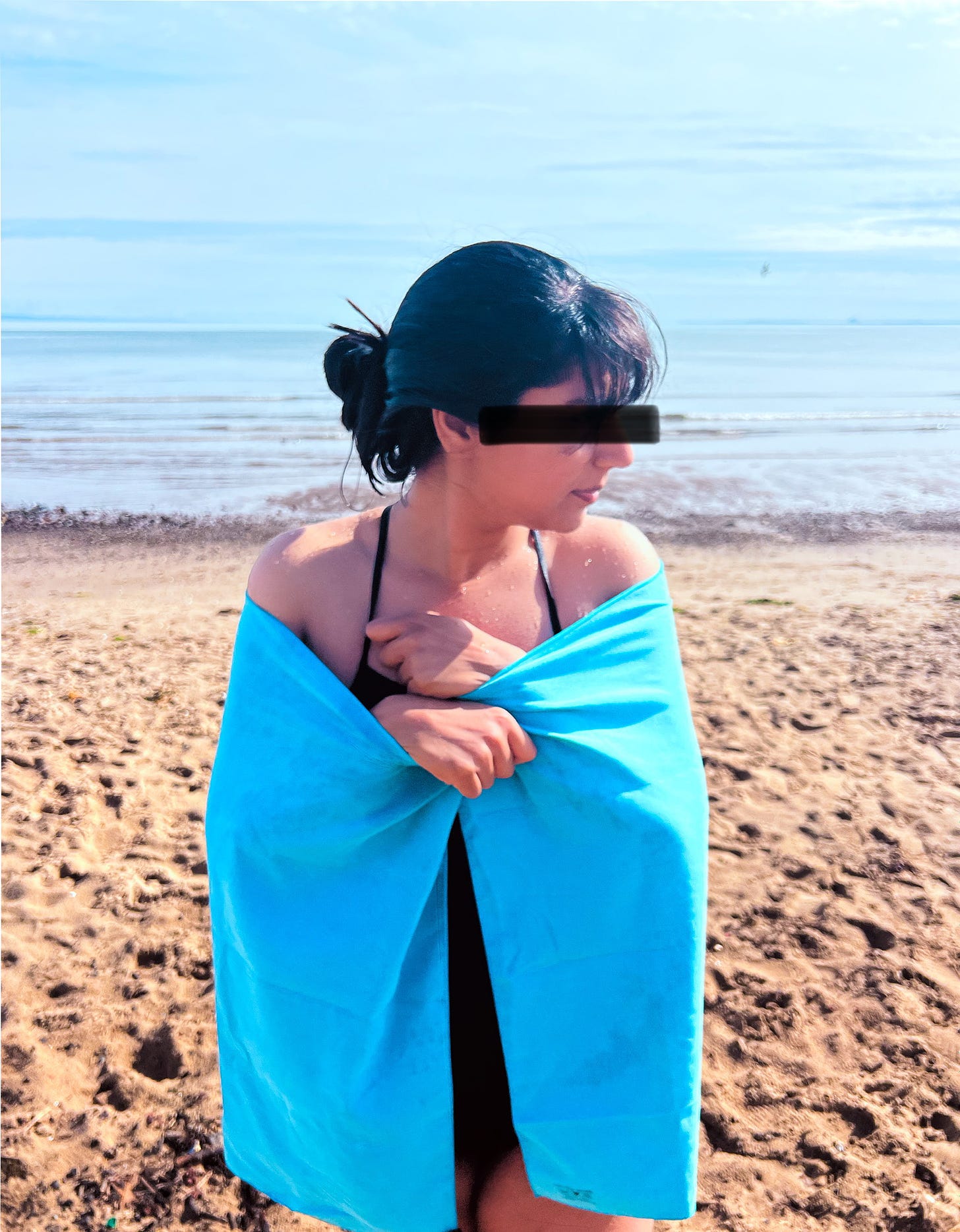 A woman stands on a beach. She is wrapping a blue towel around herself. Behind the towel is a black swimsuit. Her hair is dark and tied up. She has water droplets on her skin. The day is bright and warm looking. A black streak is drawn across her face, redacting her eyes.