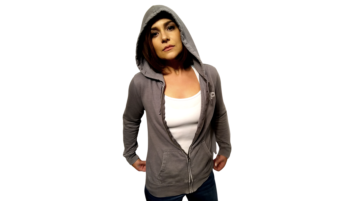 Image of a woman in a grey hooded jacket. The hood is up over her head. She has dark brown hair and is looking seriously into the camera. She wears a white tank top beneath the jacket. The background of the image is transparent and shares that of the web page which is white.
