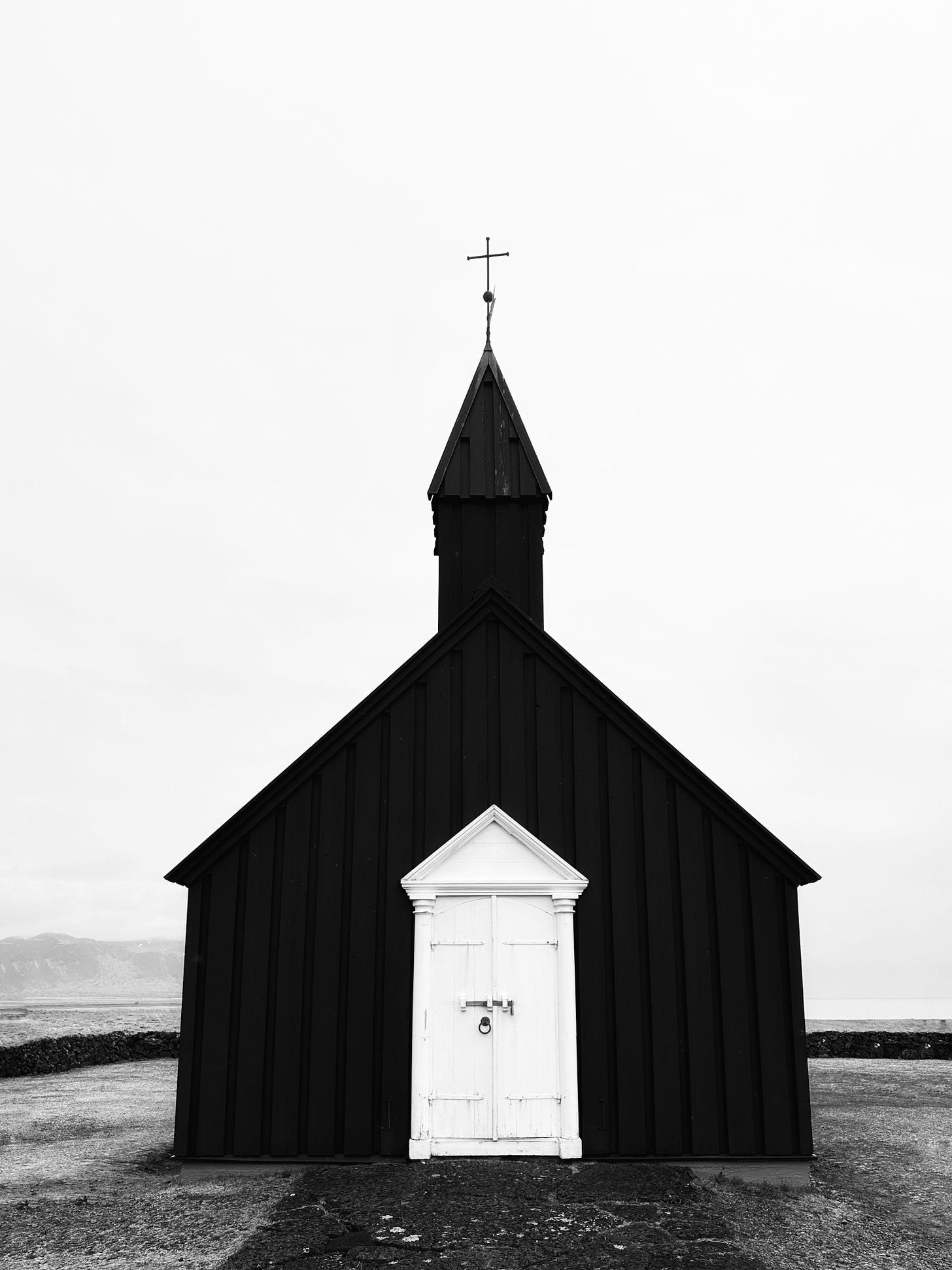 Búðakirkja - The Black Church of Budir in black and white.  This image shows the front of the church with it's weathered white door in silhouette against the light gray sky.