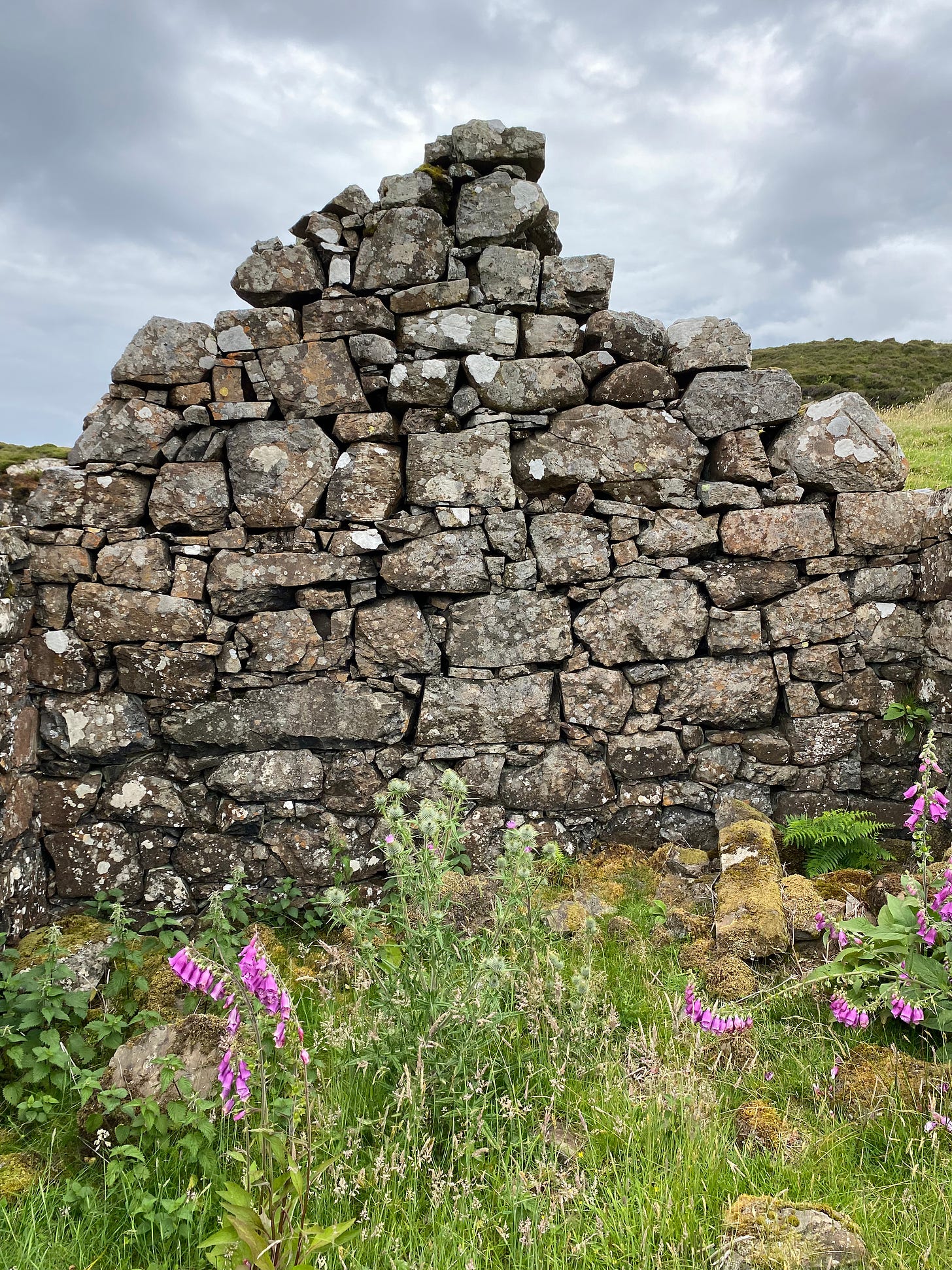 The side of a house, made out of grey uneven stones. Some of them have crumbled down and there is no roof - as it would have lilkely been a thatched roof. There is a grey sky overhead and grass and a few floors where in the home interior would have been.