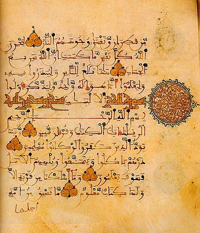 Pale yellow-orange page bearing black script in Arabic as well as leaf-shaped markers denoting the end of each ayat (phrase). At the right of the page is a large sun-shaped mark in orange and light blue.