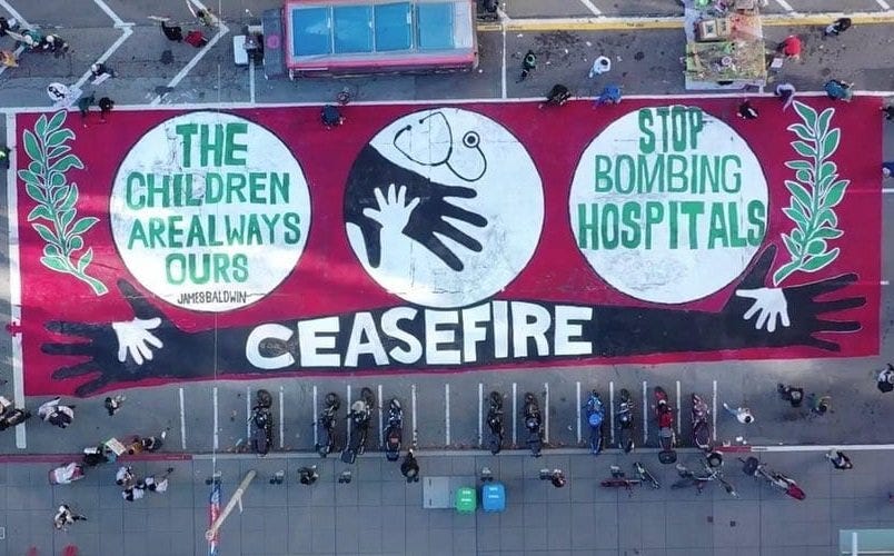 An aerial photo of a red, green, black and white mural containing three circles drawn side by side. The first circle contains the following quote by James Baldwin: “The children are always ours.” The second contains a drawing of a child’s hand resting in a larger hand with a stethoscope above. The last circle contains the words, “Stop bombing hospitals”, At the bottom of the mural is the word “Ceasefire” drawn on outstretched hands with smaller hands resting in their palms. At the sides are drawings of olive branches. People are gathered around the mural on all sides.